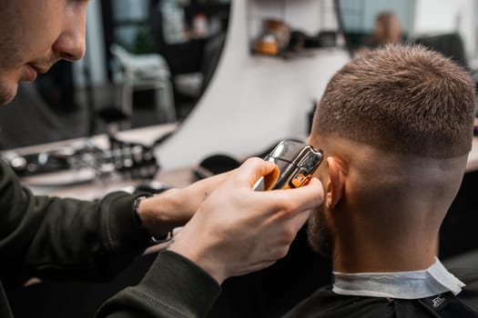 Hairstylist cuts client red hair over ears with trimmer in barbershop closeup. Master uses machine and brush doing masculine hairstyle in salon
