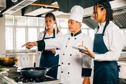 Senior Asian chef in a restaurant kitchen instructs multiracial students. Emphasizing teamwork learning note-taking. Professional education and skill-building in action. Food Edocation