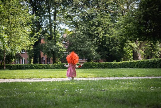 Portrait of one beautiful Caucasian little girl in a pink dress with a red Belgian flag wig on her head runs from her back across the lawn in a city park on a summer day, close-up side view with copy space on the sides.