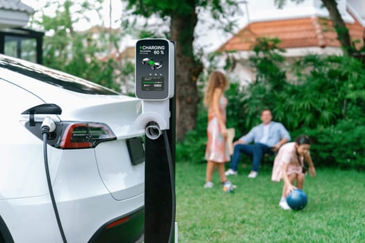 Focus electric vehicle recharge from home charging station on blur background of happy and playful family playing together. EV car using alternative and sustainable energy for better future.Synchronos