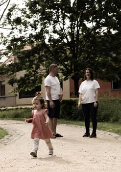 Portrait of one beautiful Caucasian baby girl in a pink dress walking and running along a path in the park with blurred parents behind her, close-up side view.