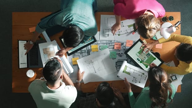 Top down aerial view of skilled civil engineer talking about building construction while standing at meeting table with project plan, architectural equipment, blueprint and color palettes. Symposium.