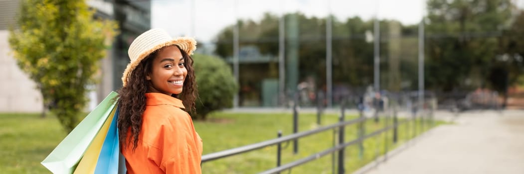 Cheerful millennial african american woman in casual and hat with many bags with purchases, looking at camera in city street. Urban walk, black friday sale, lifestyle and discount, copy space
