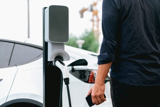 Man using smartphone online banking application to pay for electric car battery charging from EV charging station during vacation holiday road trip at national park or summer forest. Exalt