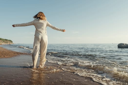 A woman stands on a sandy beach next to the ocean, gazing at the waves rolling in under a clear sky.