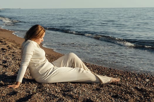 A woman sits on a sandy beach next to the ocean, gazing out at the waves crashing against the shore under a clear blue sky.