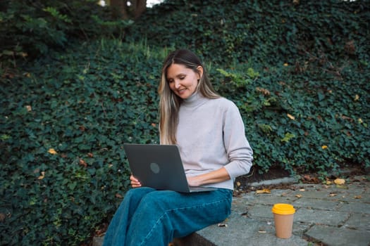 A woman is sitting on a stone ledge with a laptop and a cup of coffee. She is working or studying, as she is focused on her laptop. Concept of productivity and concentration