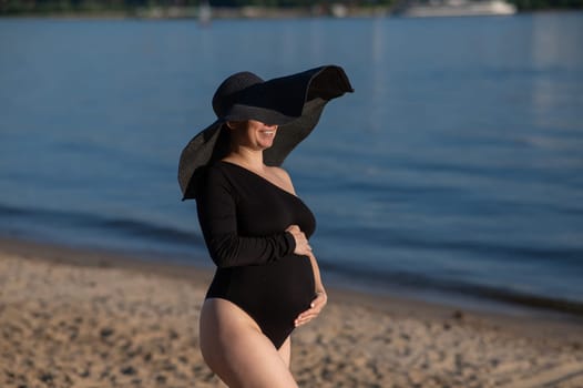 Pregnant woman in a large straw hat and black swimsuit posing on the beach