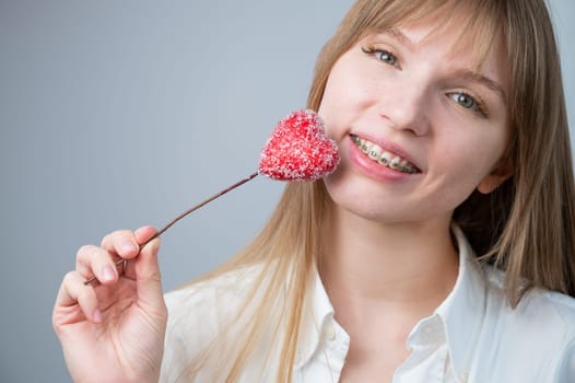 Cute woman with braces on her teeth holds a candy in the form of a heart on white background. Copy space.
