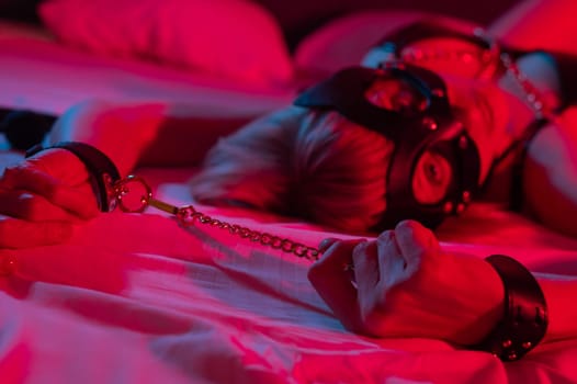 Sexy blonde woman in a leather mask and handcuffs and a collar lies in the bedroom in a red-blue neon light. Role-playing games