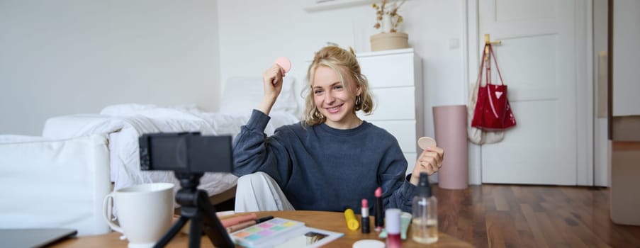 Portrait of young woman, beauty content creator, sitting in a room in front of digital camera, recording makeup tutorial vlog, showing cosmetic facial products.