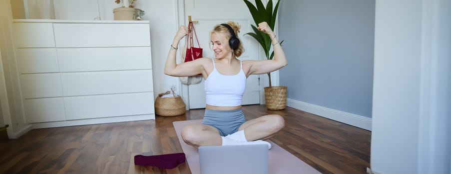Portrait of young athletic woman doing workout at home, shows her muscles, strong biceps, sits on yoga mat and wears wireless headphones.