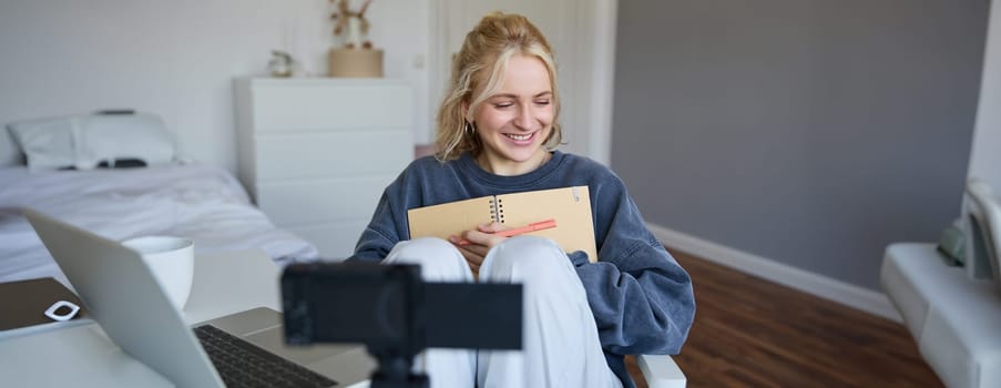 Portrait of smiling cute woman, lifestyle blogger, sits in her room with daily journal or planner, records video on digital camera, creates content for social media about daily routine.