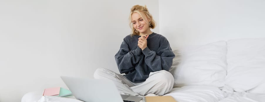 Portrait of young beautiful woman sitting on bed with laptop and notebooks, working from home, freelancing. Female student studying online, video chats, connects to lesson remotely.