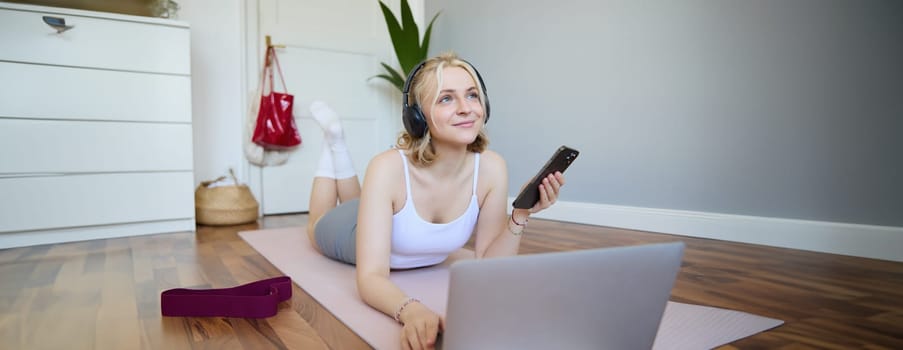 Portrait of happy, smiling sporty woman, doing workout at home, using laptop to watch workout videos in headphones, checking sports app on mobile phone.