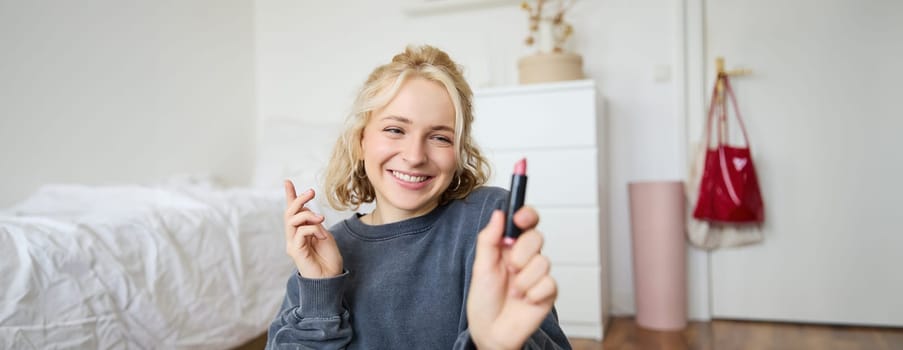 Portrait of young social media influencer, woman recording a video with beauty products, showing her makeup on camera, holding lipstick and smiling, sitting on floor.