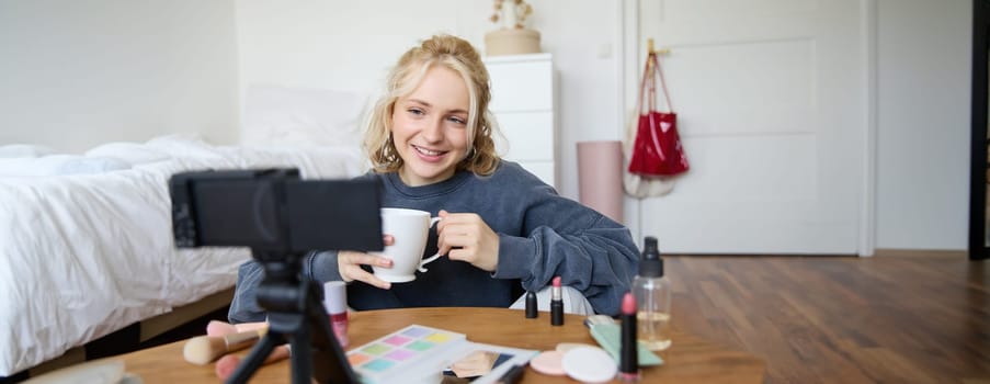 Blogging, modern technology, e-commerce and people concept. Happy smiling woman beauty video blogger holding cup of tea applying makeup while recording beauty tutorial at home for her video blog.
