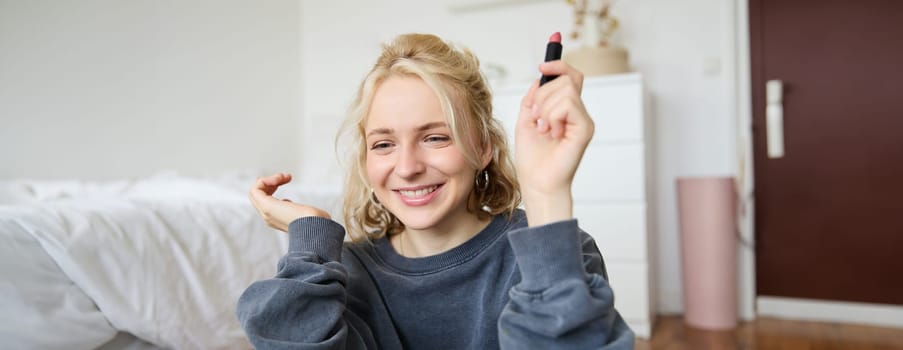 Portrait of young social media influencer, woman recording a video with beauty products, showing her makeup on camera, holding lipstick and smiling, sitting on floor.