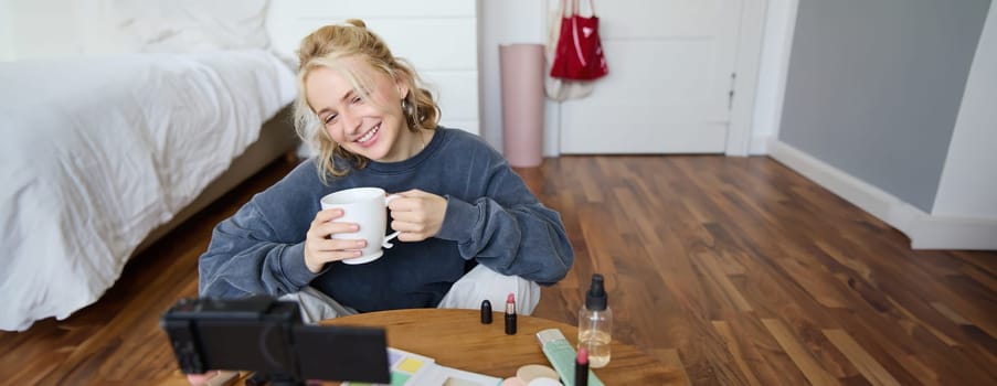 Young woman smiling, recording lifestyle vlog on her digital camera, holding cup of tea while talking, has makeup on coffee table, doing makeup tutorial for social media from her room.