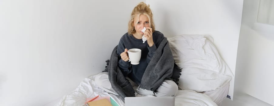 Portrait of young woman sitting at home sick, feeling unwell, drinking tea, spending time in bed, has runny nose, working on laptop.