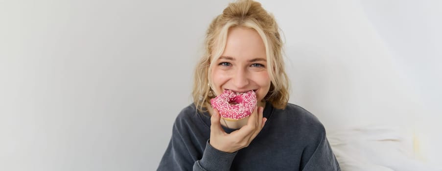 Portrait of cute blond girl holding pink doughnut with sprinkles on top, showing her favourite food.