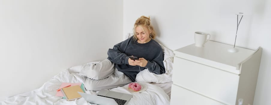 Lifestyle and people concept. Young beautiful woman, staying at home, lying in bed with laptop and smartphone, eating doughnut, enjoying free time, spending weekend at home, watching movie online.