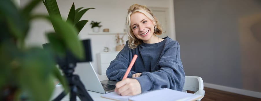 Portrait of young woman, lifestyle blogger, recording video of herself, making notes, writing in journal, sitting in front of laptop in a room and studying.