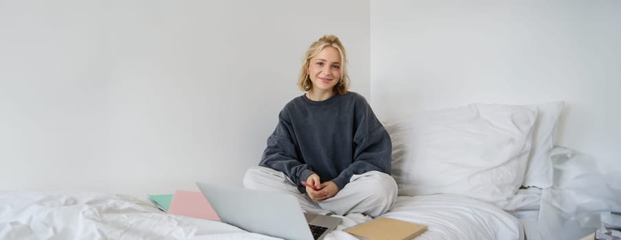 Portrait of female student, woman sits on bed with laptop and notebooks, studying online, remote education concept. Girl freelancer working from her bedroom.