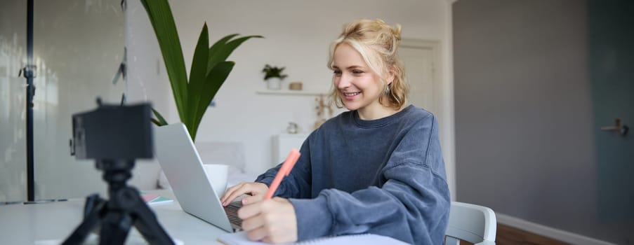 Portrait of young blond smiling woman, studying at home, remote education concept, connects to online course or lesson, writing in notebook.