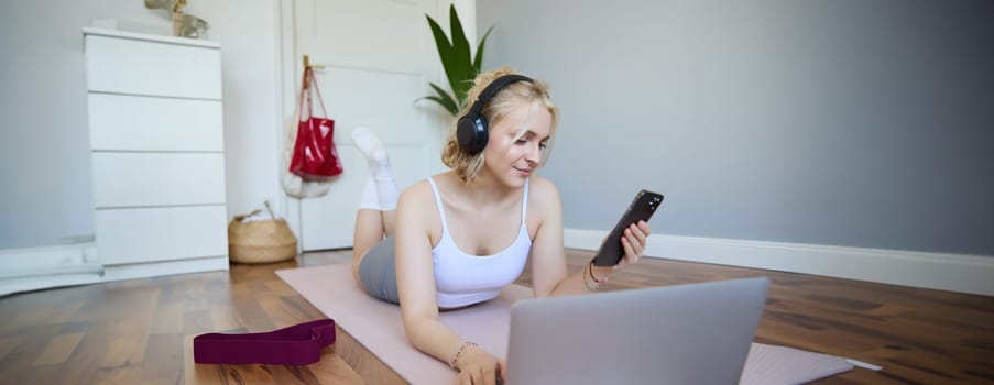 Portrait of happy, smiling sporty woman, doing workout at home, using laptop to watch workout videos in headphones, checking sports app on mobile phone.