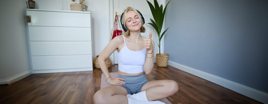 Portrait of happy, fit and healthy young woman sitting on yoga mat, wearing wireless headphones, showing thumbs up with satisfied face, pleased with workout training at home.