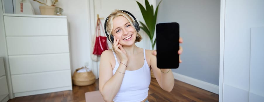 Portrait of smiling, cheerful young woman workout at home and showing smartphone screen, wearing headphones, recommending mobile app. Lifestyle concept