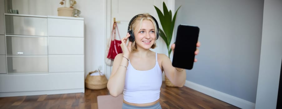 Portrait of fit and healthy woman workout at home, wearing headphones, showing smartphone screen, empty dark display. Wellbeing and lifestyle concept