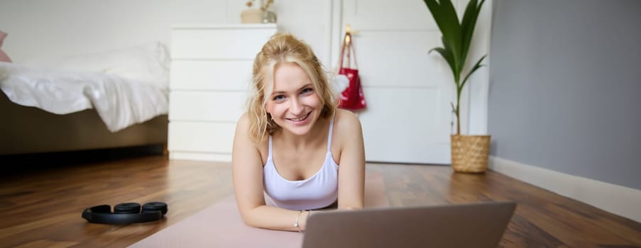 Portrait of beautiful blond woman looking at fitness video tutorials on laptop, lying on rubber yoga mat, following workout instructions online.