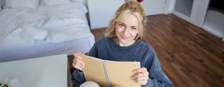 Portrait of smiling, charismatic young woman, writing down notes, making plans and putting it in planner, holding journal, sitting in bedroom and looking happy at camera.