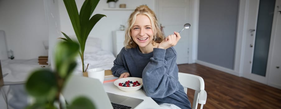 Image of laughing woman sitting in front of laptop in her room, eating breakfast, holding spoon and bowl in hand, watching videos online while having a snack.