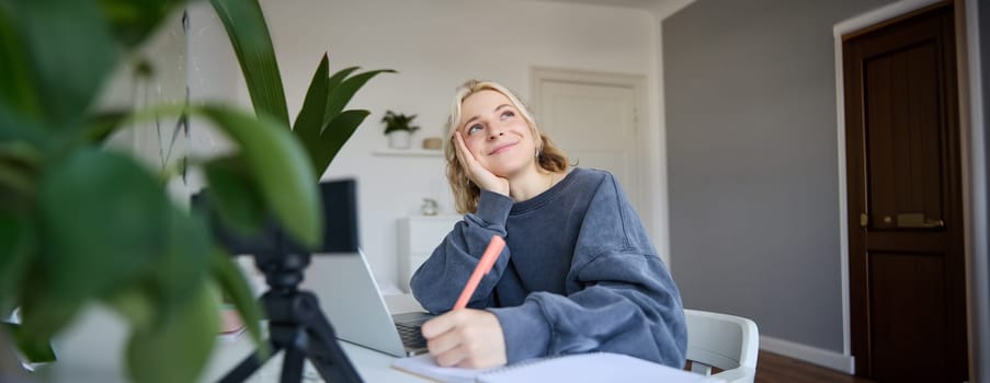 Portrait of young woman, lifestyle blogger, recording video of herself, making notes, writing in journal, sitting in front of laptop in a room and studying.