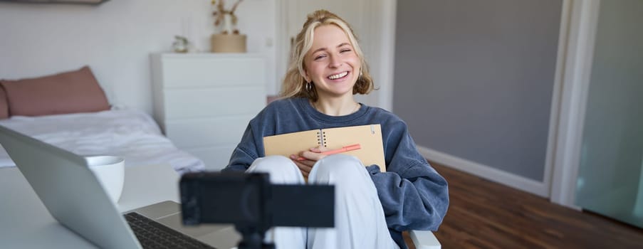 Portrait of smiling, charismatic young woman, content creator, records video on digital camera, laughing and looking happy, using journal, writing in notebook.