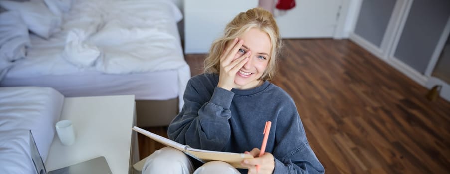 Portrait of smiling, charismatic young woman, writing down notes, making plans and putting it in planner, holding journal, sitting in bedroom and looking happy at camera.
