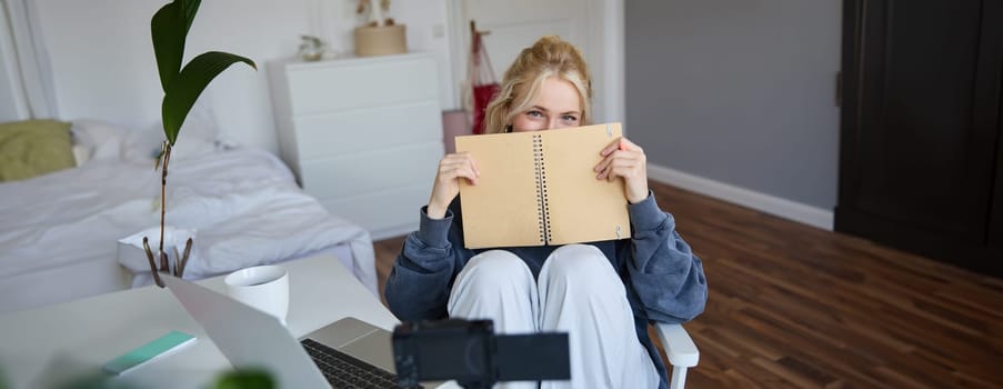 Portrait of cute blond woman, sits in front of digital camera and laptop in her room, covers face with notebook journal, records video of herself for lifestyle vlog.