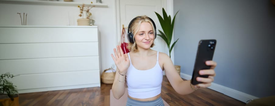 Portrait of woman waving hand at smartphone, wearing wireless headphones, working out and recording video for social media account about fitness.