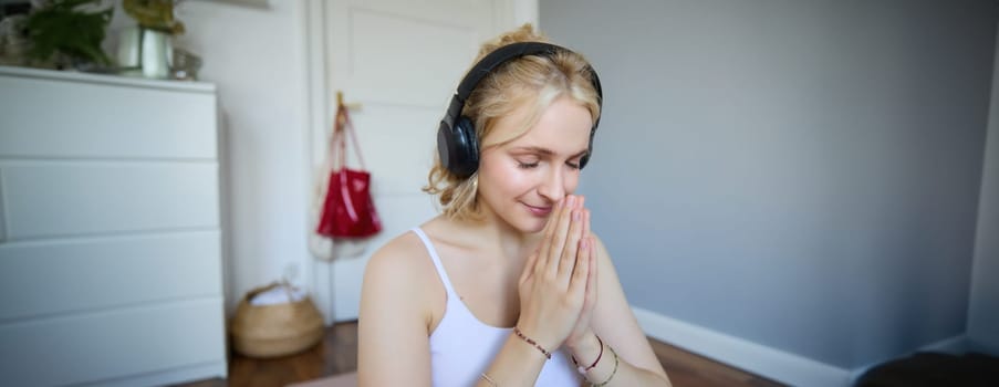 Portrait of young relaxed woman, sits in room in headphones, clasp hands together, meditated, listens to yoga music.