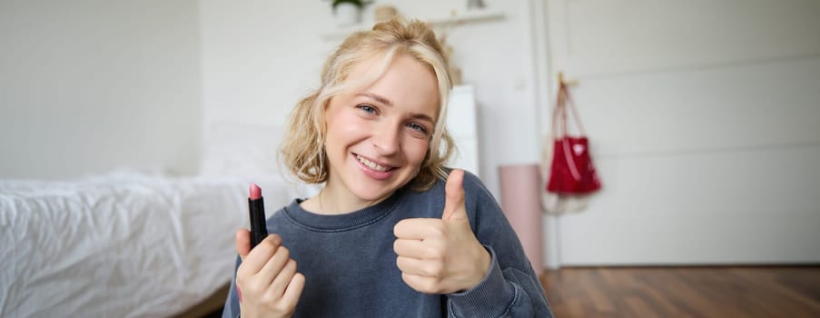 Cute smiling girl shows lipstick and thumbs up hand sign, recommending beauty product, records content for social media account, lifestyle vlog.