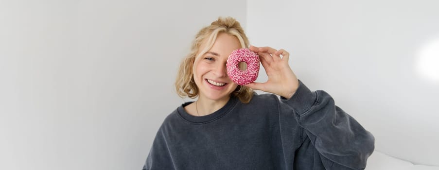 Close up portrait of happy, cute blond woman, holding doughnut, eating sweet, delicious comfort food, showing dessert at camera. Food concept