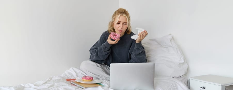 Portrait of woman watching sad show on laptop, eating doughnut and wiping tears with napkin, sitting on bed, spending time at home while being upset.