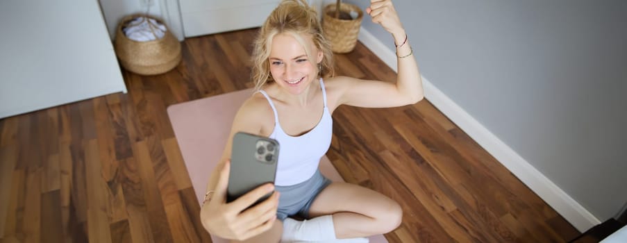 Portrait of blond young woman, fitness training, sitting on yoga mat, taking selfies during workout at home, showing muscles, flexing biceps.