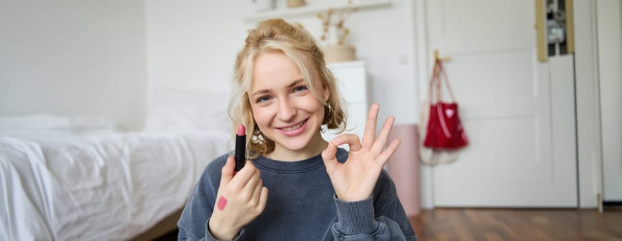 Portrait of young blond woman, content creator, recording video for social media about makeup, showing okay hand sign and lipstick, recommending good cosmetic product.
