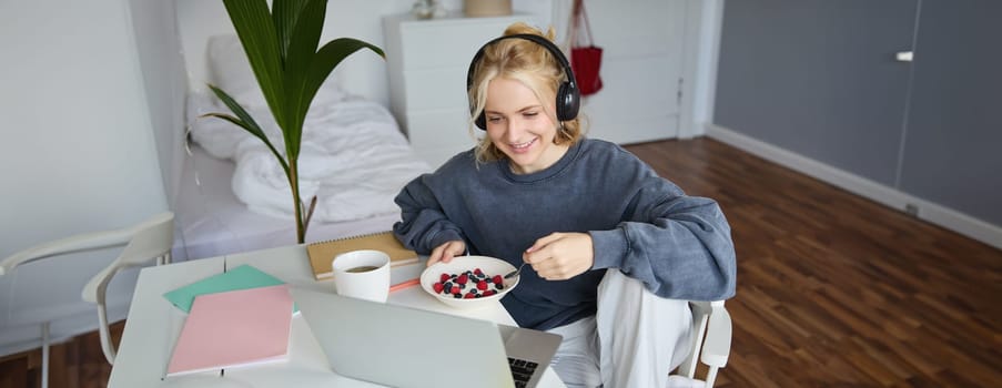 Portrait of happy young blond woman, sitting in a room, watching movie on laptop and eating healthy breakfast, drinking tea, resting on weekend.
