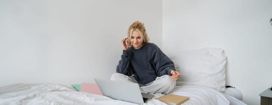 Portrait of female student, woman sits on bed with laptop and notebooks, studying online, remote education concept. Girl freelancer working from her bedroom.