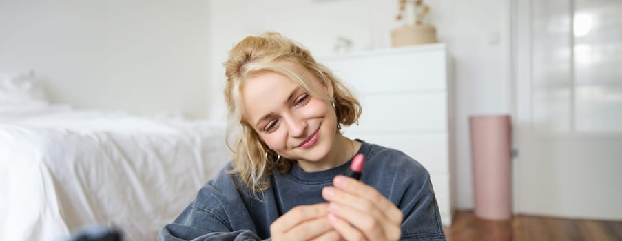 Portrait of smiling beautiful woman in her room, sitting and showing lipstick, recommending favourite beauty product, content maker recording a video of herself for social media blog.